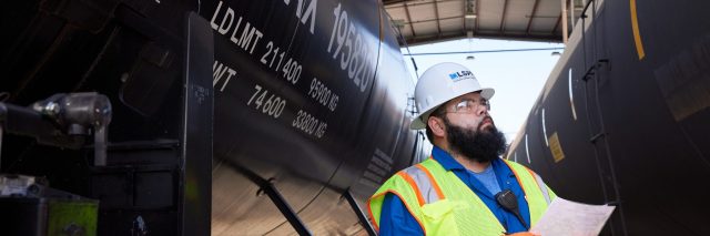 "Industrial worker wearing a hard hat and high-visibility safety vest reviewing paperwork between two black tanker railcars inside a covered rail loading facility."