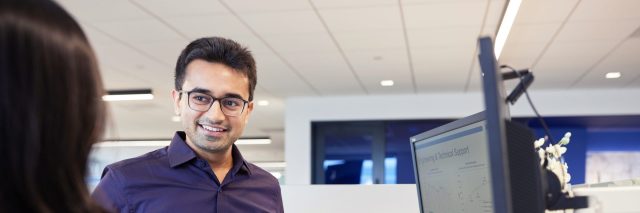"Two professionals standing at a sit-stand workstation reviewing information on a desktop monitor in a modern office environment."